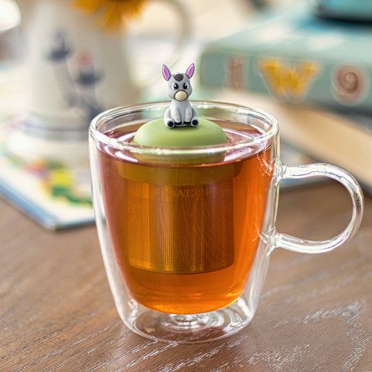 grey donkey tea infuser for loose leaf tea with a fine mesh stainless steel tea strainer that reads FLOAT2INFUSE on a green base floating in a glass tea mug 
with books in the background