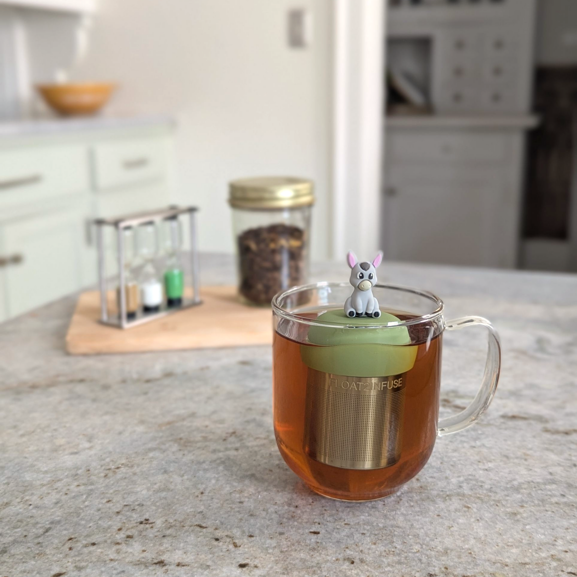 grey donkey tea infuser for loose leaf tea with a fine mesh stainless steel tea strainer that reads FLOAT2INFUSE on a green base floating in a glass tea mug 
with loose leaf tea in the background