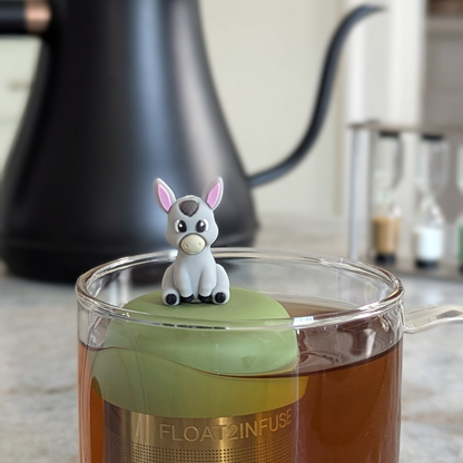 grey donkey tea infuser for loose leaf tea with a fine mesh stainless steel tea strainer that reads FLOAT2INFUSE on a green base floating in a glass tea mug 
with a tea kettle in the background