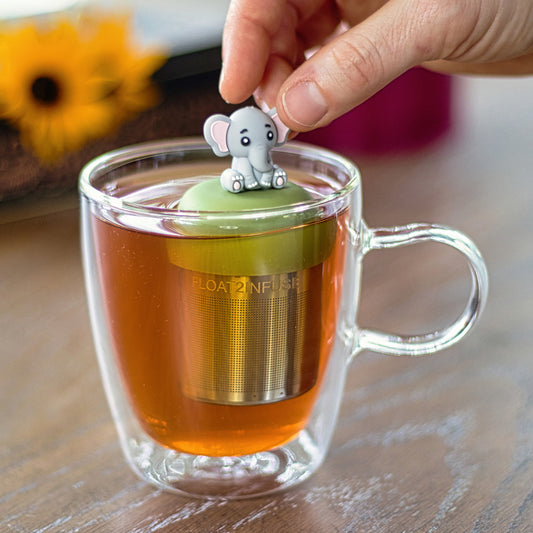 cute grey elephant tea infuser for loose leaf tea with a fine mesh stainless steel tea strainer that reads FLOAT2INFUSE on a green base  floating in a glass tea mug 
with flowers in the background