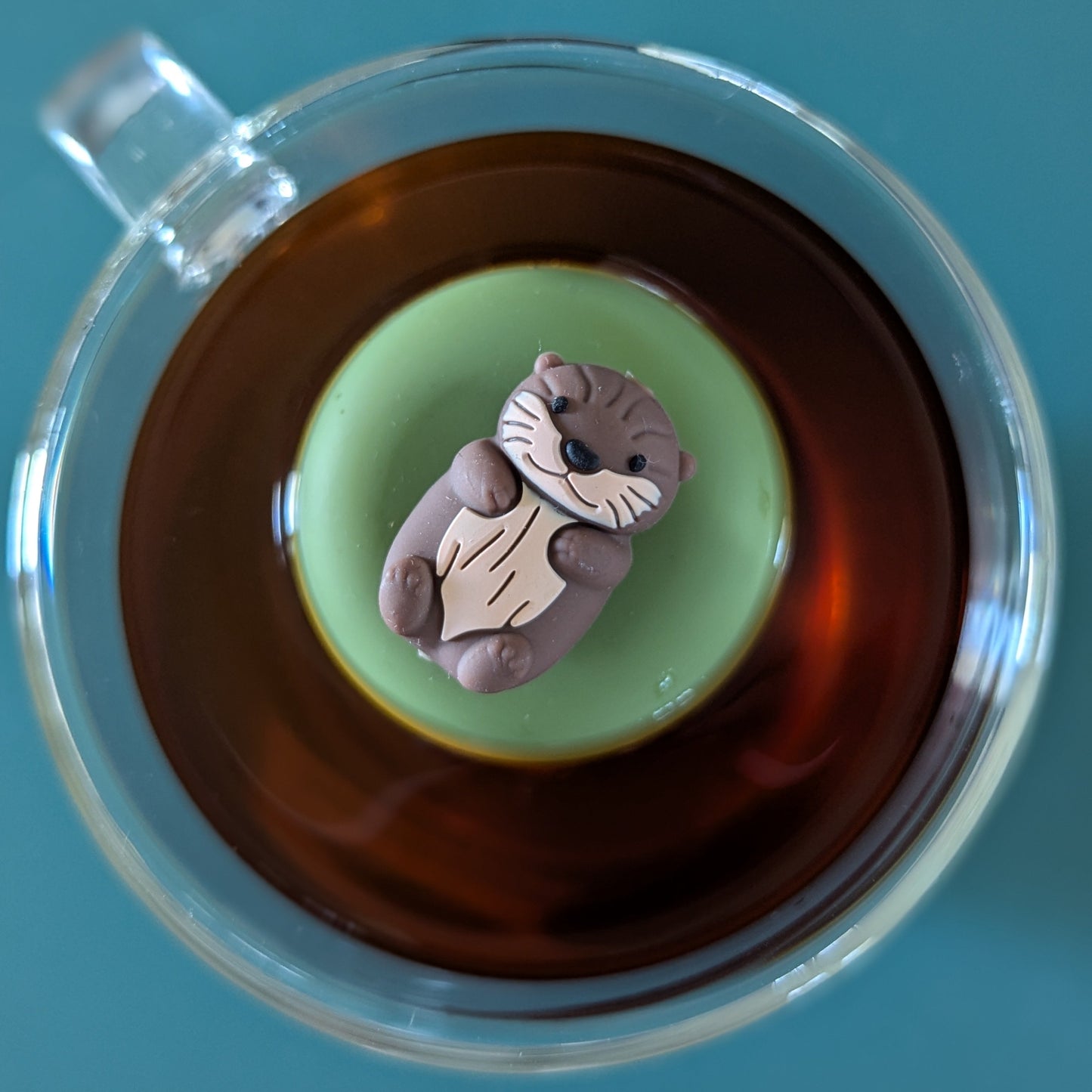 top view of a brown otter tea infuser for loose leaf tea with a fine mesh stainless steel tea strainer that reads FLOAT2INFUSE on a green base floating in a glass tea mug
