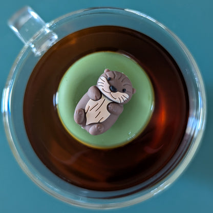 top view of a brown otter tea infuser for loose leaf tea with a fine mesh stainless steel tea strainer that reads FLOAT2INFUSE on a green base floating in a glass tea mug
