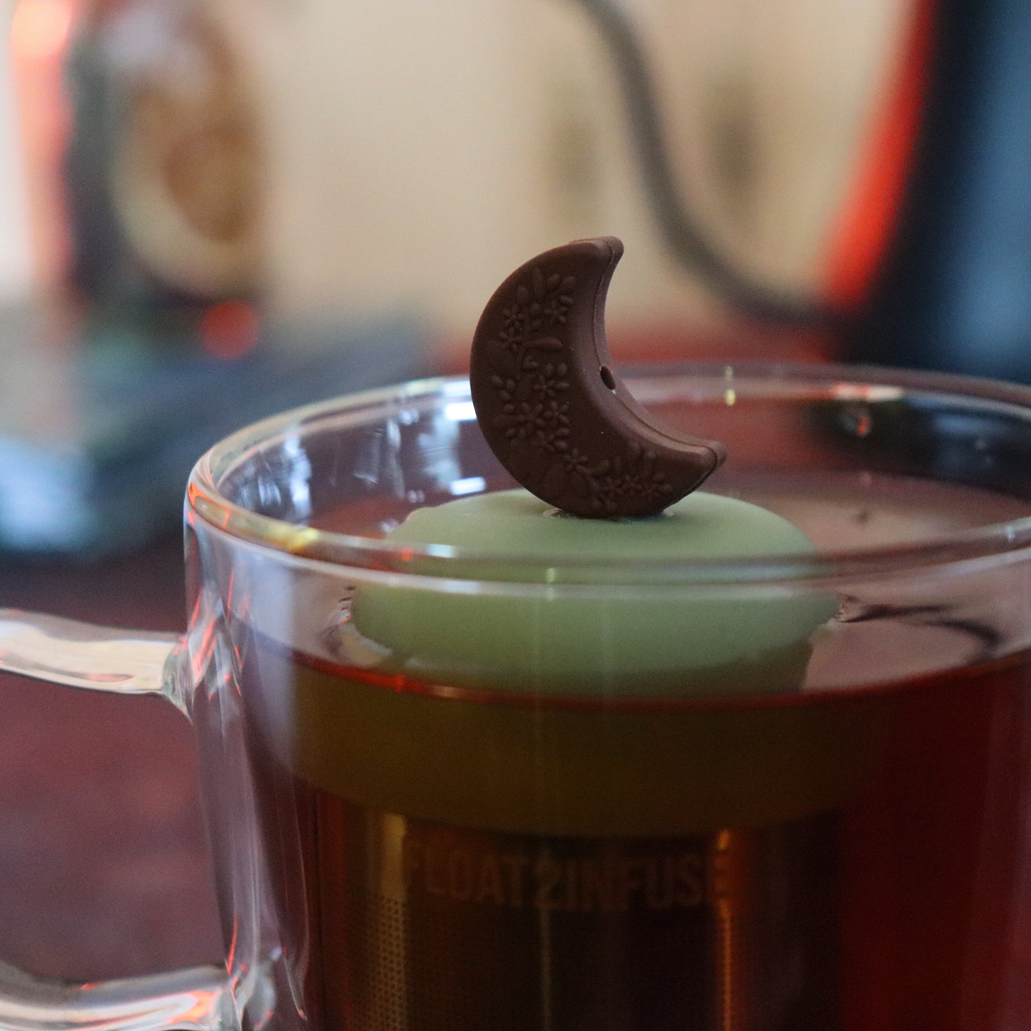 brown crescent floral moon tea infuser for loose leaf tea  with a fine mesh stainless steel tea strainer that reads FLOAT2INFUSE on a green base floating in a glass tea mug with tea kettle in the background