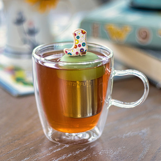 white cowboy boot with multicolored floral pattern tea infuser for loose leaf tea with a fine mesh stainless steel tea strainer that reads FLOAT2INFUSE on a green base  floating in a glass tea mug with books in the background