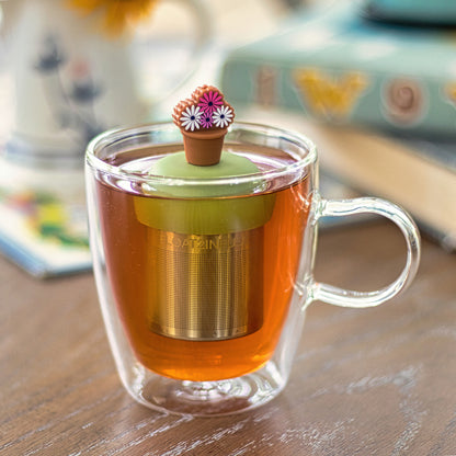 brown flower pot with pink and white flowers tea infuser for loose leaf tea with a fine mesh stainless steel tea strainer that reads FLOAT2INFUSE on a green base floating in a glass tea mug 
with books in the background