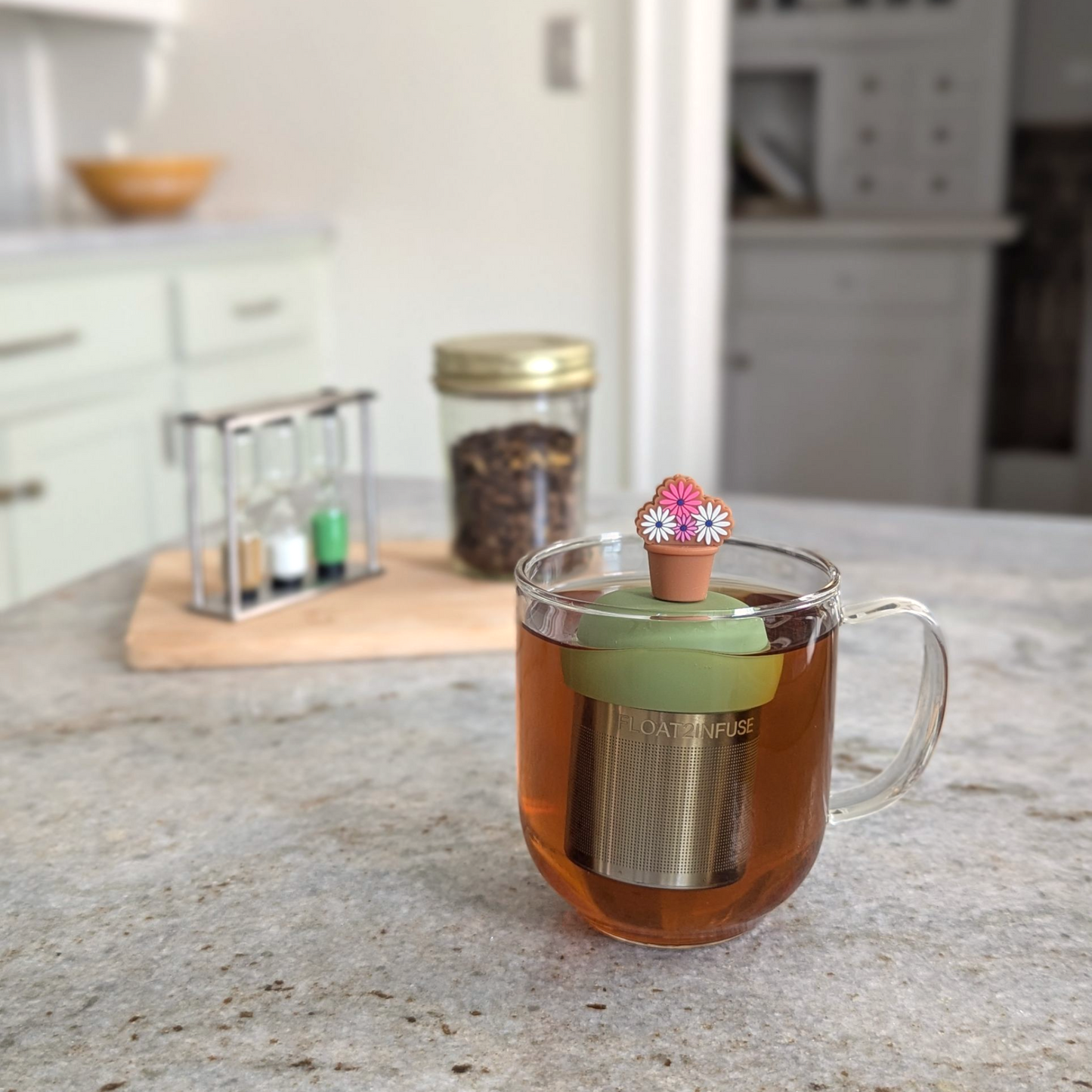 brown flower pot with pink and white flowers tea infuser for loose leaf tea with a fine mesh stainless steel tea strainer that reads FLOAT2INFUSE on a green base floating in a glass tea mug 
with loose leaf tea in the background