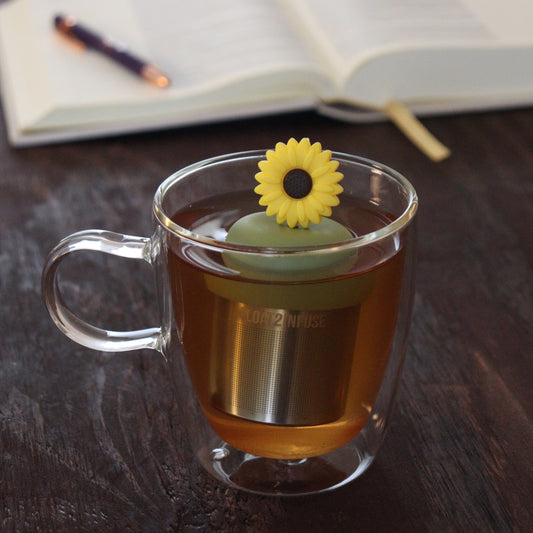 yellow sunflower tea infuser for loose leaf tea on a green base with a fine mesh stainless steel tea infuser that reads FLOAT2INFUSE floating in a glass tea mug with notebook and pen in the background