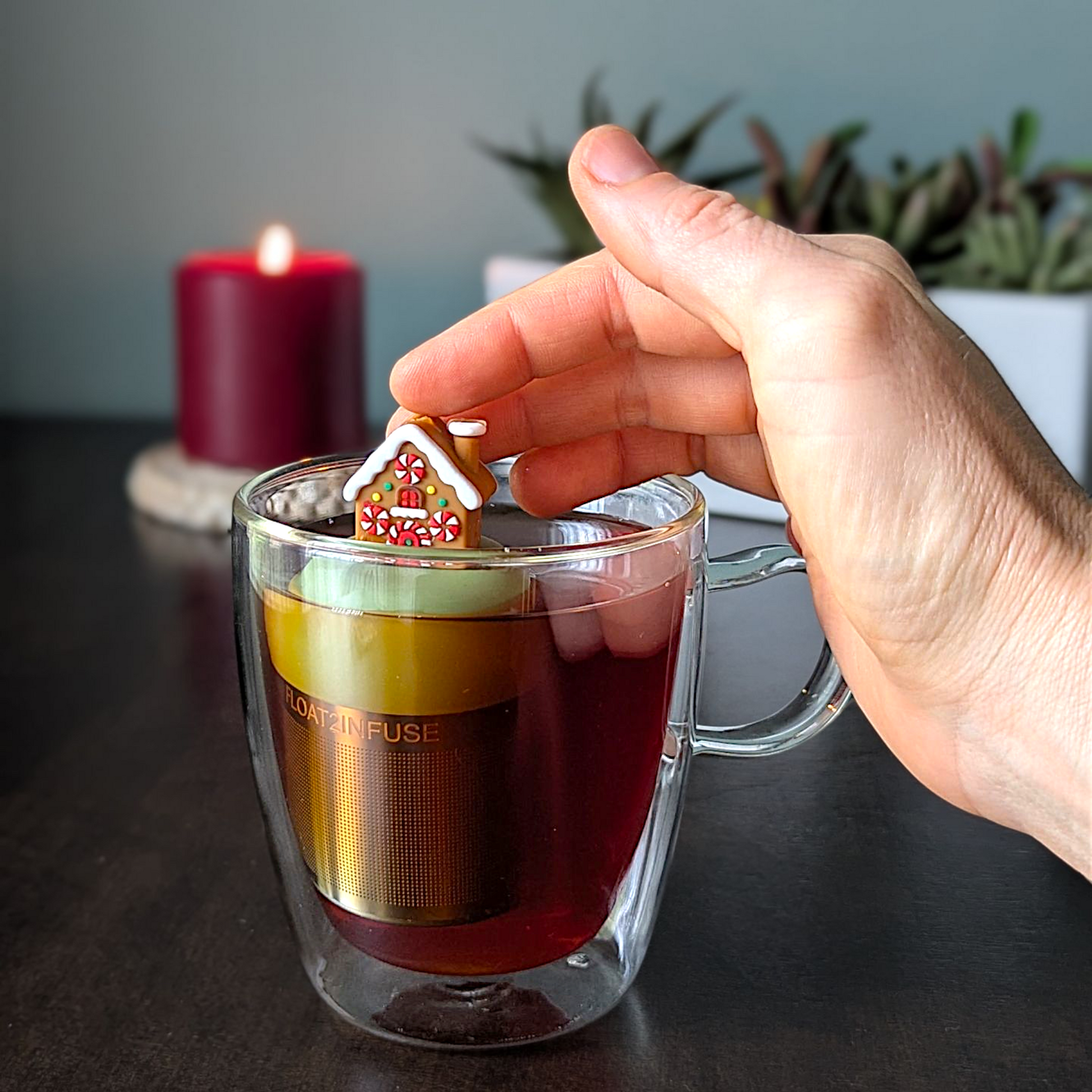 Brown gingerbread house with peppermint candy windows and snow on the roof tea infuser for loose leaf tea with a fine mesh stainless steel tea strainer that reads FLOAT2INFUSE on a green base with a lit candle and plants in the background

