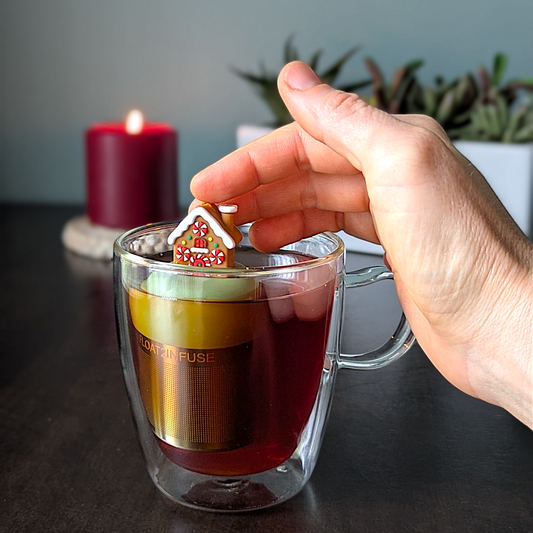 Brown gingerbread house with peppermint candy windows and snow on the roof tea infuser for loose leaf tea with a fine mesh stainless steel tea strainer that reads FLOAT2INFUSE on a green base with a lit candle and plants in the background
