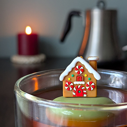 Brown gingerbread house with peppermint candy windows and snow on the roof tea infuser for loose leaf tea with a fine mesh stainless steel tea strainer that reads FLOAT2INFUSE on a green base with a lit candle and a tea kettle in the background
