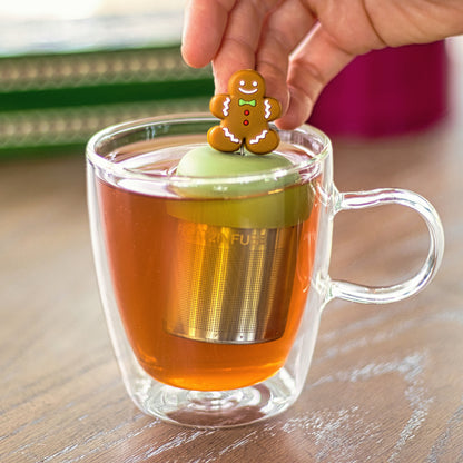 smiling gingerbread man with green bowtie, red buttons and white accents tea infuser for loose leaf tea with a fine mesh stainless steel tea strainer that reads FLOAT2INFUSE on a green base floating in a glass tea mug with books in the background