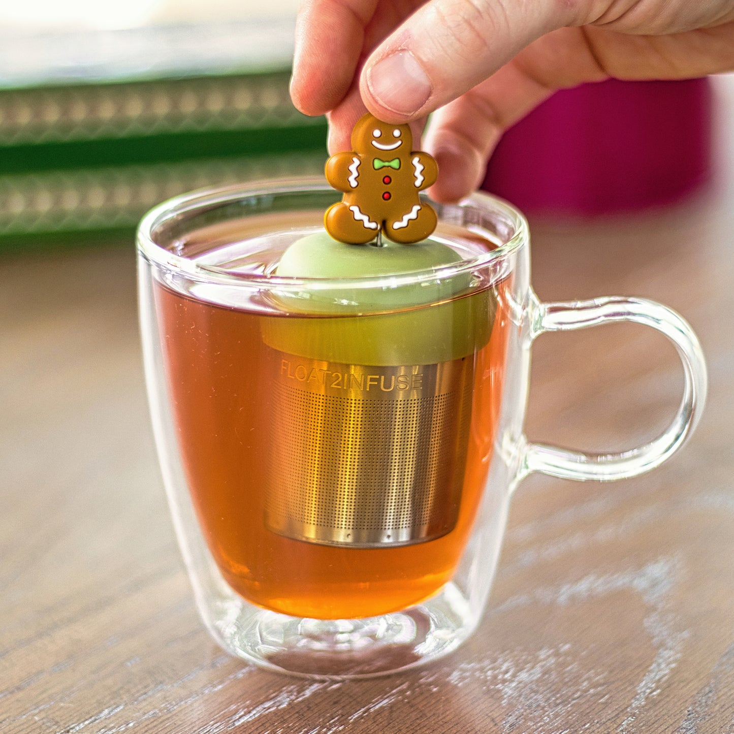 smiling gingerbread man with green bowtie, red buttons and white accents tea infuser for loose leaf tea with a fine mesh stainless steel tea strainer that reads FLOAT2INFUSE on a green base floating in a glass tea mug with books in the background