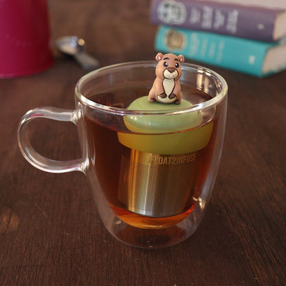 Sassy guinea pig tea infuser for loose leaf tea with a fine mesh stainless steel tea strainer 
that reads FLOAT2INFUSE on a green base with a tea canister and books in the background