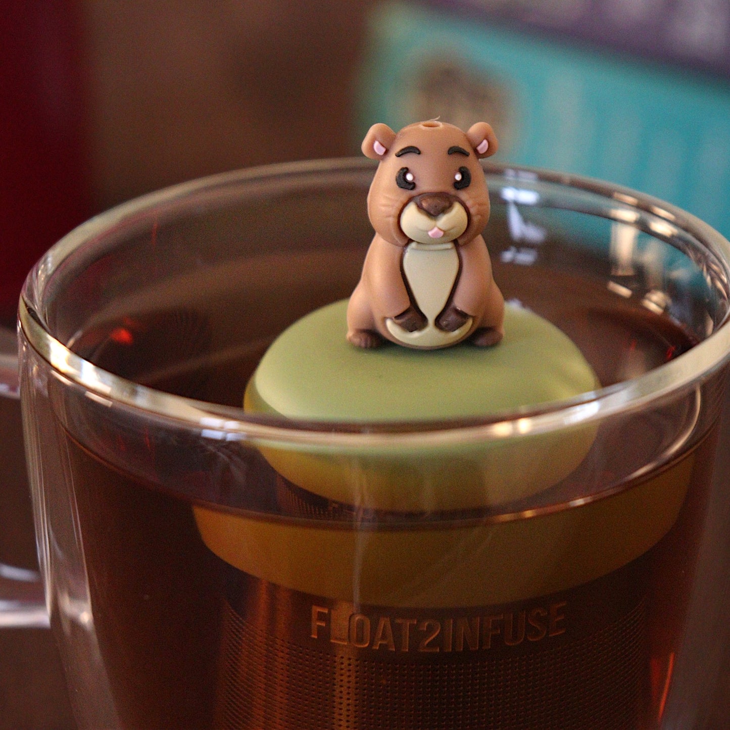 Sassy guinea pig tea infuser for loose leaf tea with a fine mesh stainless steel tea strainer 
that reads FLOAT2INFUSE on a green base with a tea canister and books in the background