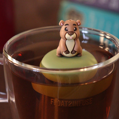 Sassy guinea pig tea infuser for loose leaf tea with a fine mesh stainless steel tea strainer 
that reads FLOAT2INFUSE on a green base with a tea canister and books in the background