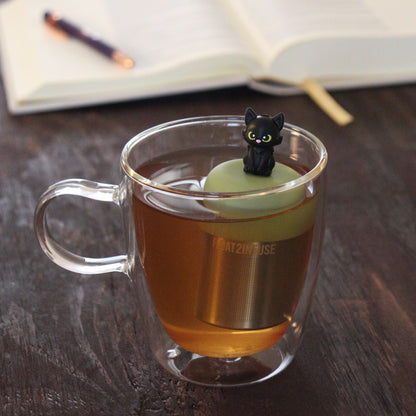 black cat animal tea infuser for loose leaf tea  with a fine mesh stainless steel tea infuser that reads FLOAT2INFUSE on a green base floating in a glass tea mug with notebook and pen in the background