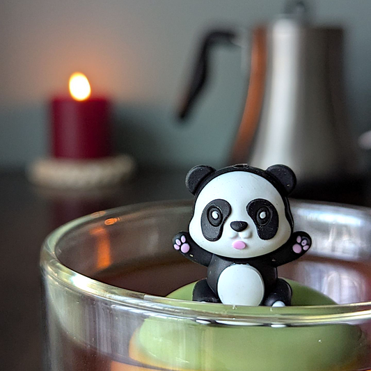 Black and white panda with outstretched arms tea  infuser for loose leaf tea with a fine mesh stainless steel tea strainer that reads FLOAT2INFUSE on a green base  with a lit candle and a tea kettle in the background