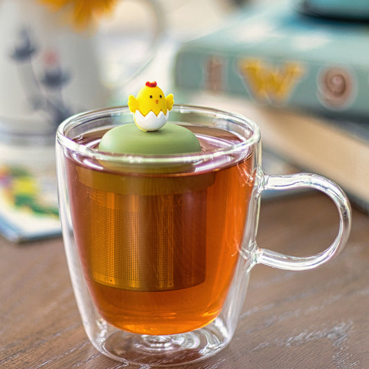 yellow chick animal hatching from white egg tea infuser for loose leaf tea with a fine mesh stainless steel tea strainer that reads FLOAT2INFUSE on a green base  floating in a glass tea mug with books in the background