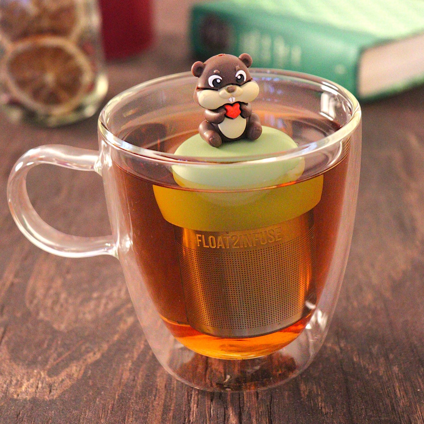 beaver animal holding a red heart  tea infuser for loose leaf tea with a fine mesh stainless steel tea infuser that reads FLOAT2INFUSE on a green base  floating in a glass tea mug with  loose leaf tea and books in the background