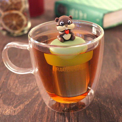 beaver animal holding a red heart  tea infuser for loose leaf tea with a fine mesh stainless steel tea infuser that reads FLOAT2INFUSE on a green base  floating in a glass tea mug with  loose leaf tea and books in the background