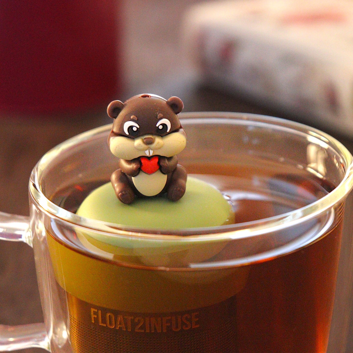 beaver animal holding a red heart  tea infuser for loose leaf tea with a fine mesh stainless steel tea infuser that reads FLOAT2INFUSE on a green base  floating in a glass tea mug with  loose leaf tea and books in the background