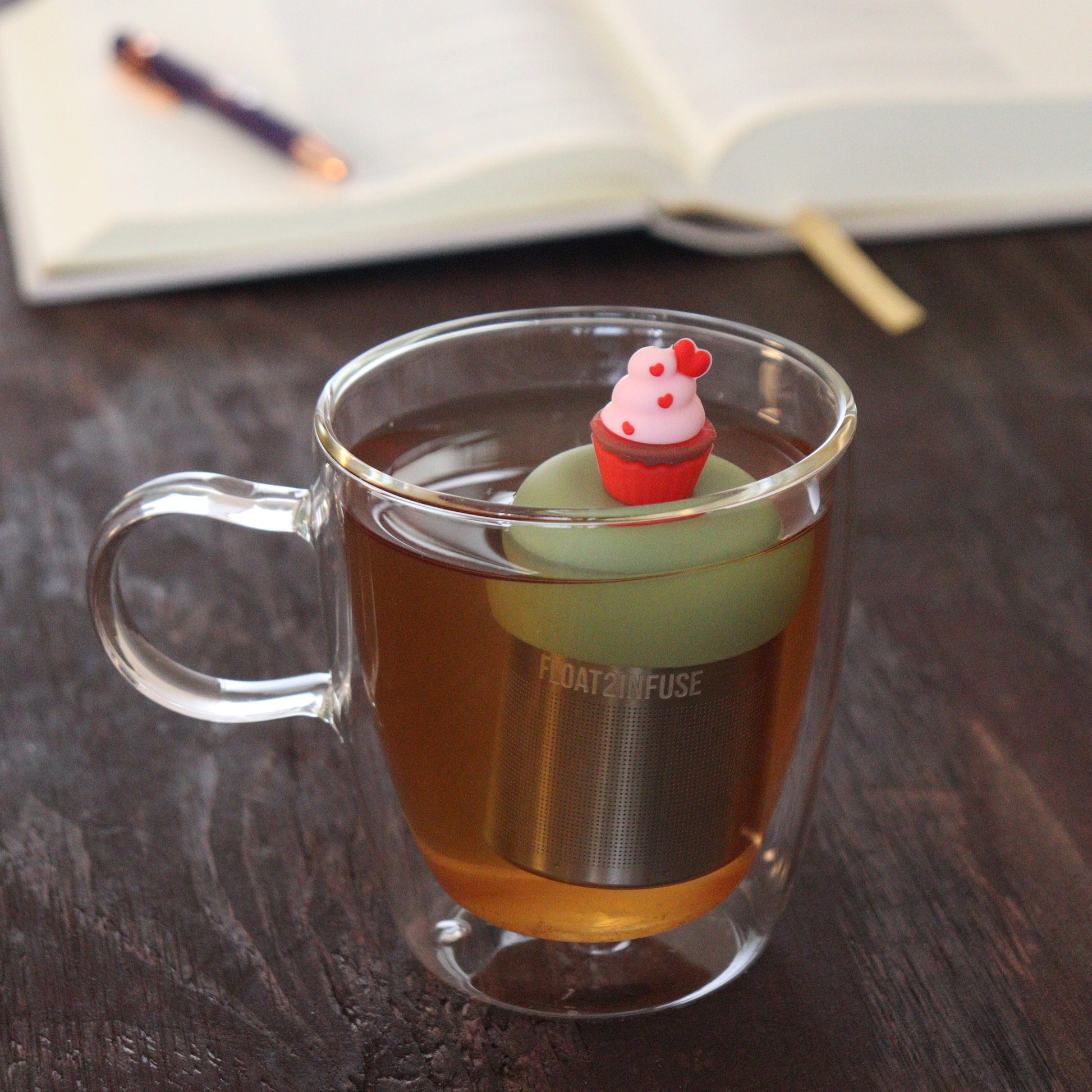 red and pink cupcake with hearts tea infuser for loose leaf tea  with a fine mesh stainless steel tea infuser that reads FLOAT2INFUSE on a green base floating in a glass tea mug with notebook and pen in the background