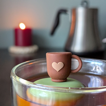 Brown tea mug with a beige heart tea infuser for loose leaf tea with a fine mesh stainless steel tea strainer that reads FLOAT2INFUSE on a green base  with a lit candle and a tea kettle in the background