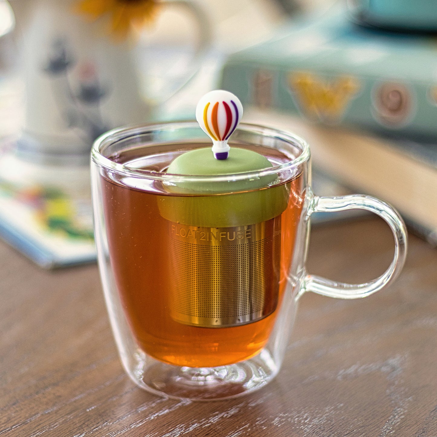 hot air ballon with rainbow stripes and purple basket tea infuser for loose leaf tea with a fine mesh stainless steel tea strainer that reads FLOAT2INFUSE on a green base floating in a glass tea mug 
with books in the background
