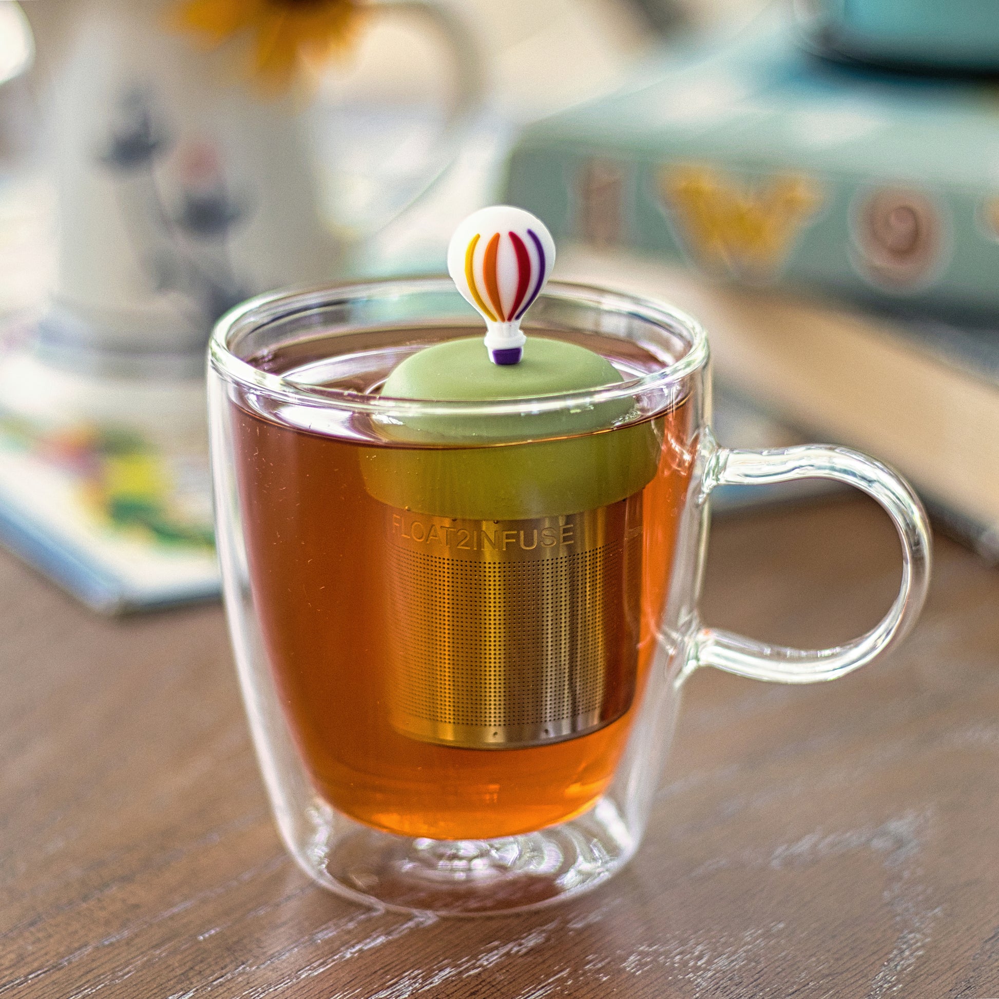 hot air ballon with rainbow stripes and purple basket tea infuser for loose leaf tea with a fine mesh stainless steel tea strainer that reads FLOAT2INFUSE on a green base floating in a glass tea mug 
with books in the background
