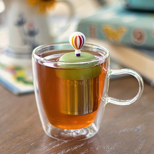 hot air ballon with rainbow stripes and purple basket tea infuser for loose leaf tea with a fine mesh stainless steel tea strainer that reads FLOAT2INFUSE on a green base floating in a glass tea mug 
with books in the background