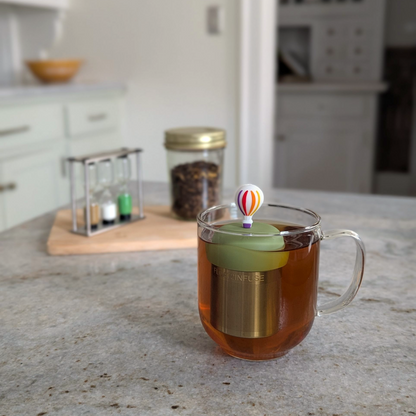hot air ballon with rainbow stripes and purple basket tea infuser for loose leaf tea with a fine mesh stainless steel tea strainer that reads FLOAT2INFUSE on a green base floating in a glass tea mug 
with loose leaf tea in the background