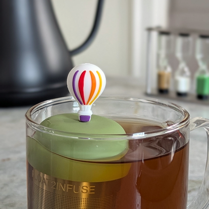 hot air ballon with rainbow stripes and purple basket tea infuser for loose leaf tea with a fine mesh stainless steel tea strainer that reads FLOAT2INFUSE on a green base floating in a glass tea mug 
with a tea kettle in the background