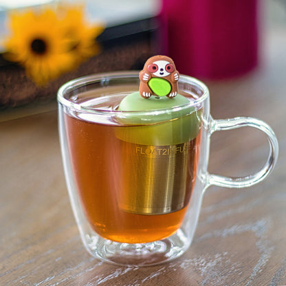 brown sloth animal holding a green leaf infuser for loose leaf tea with a fine mesh stainless steel tea strainer that reads FLOAT2INFUSE on a green base  floating in a glass tea mug 
with flowers in the background
