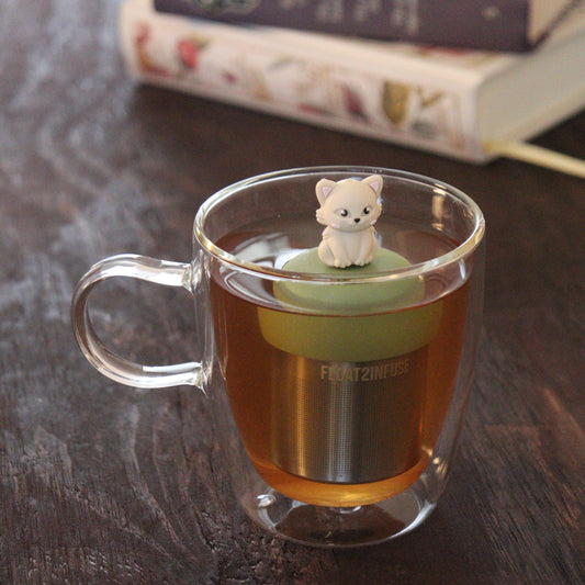 ivory cat animal tea infuser for loose leaf tea on a green base with a fine mesh stainless steel tea infuser that reads FLOAT2INFUSE floating in a glass tea mug with books in the background