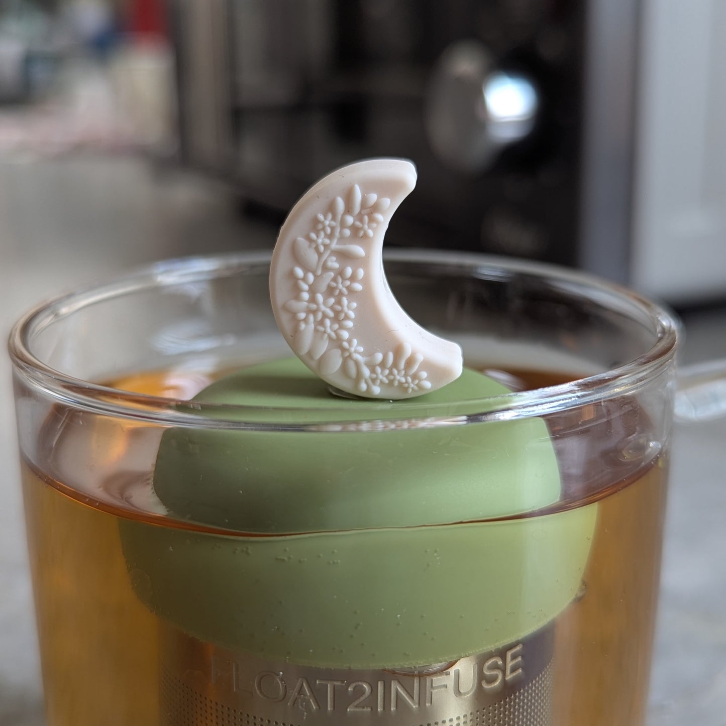 ivory crescent moon with raised floral pattern tea infuser  for loose leaf tea with fine mesh stainless steel tea strainer that reads FLOAT2INFUSE on a green base floating in a glass tea mug