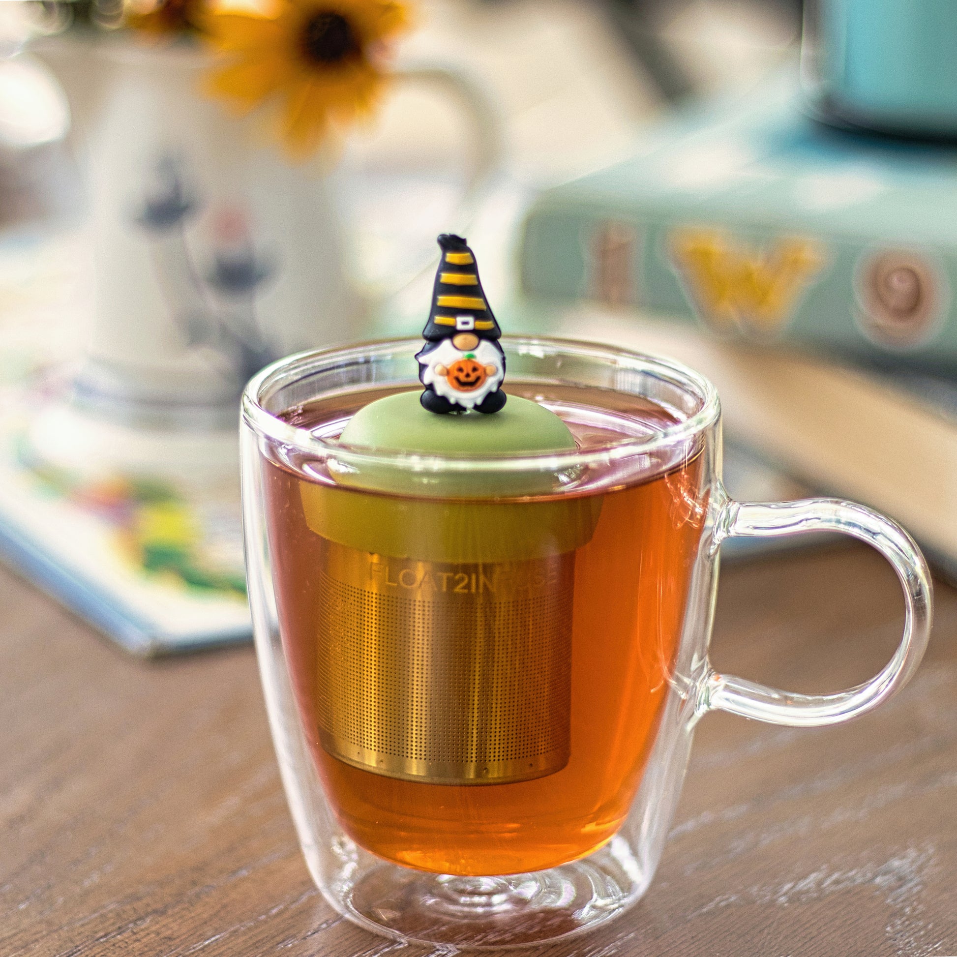 gnome with long white beard wearing black with yellow stripes and holding a jack-o-lantern pumpkin tea infuser for loose leaf tea with a fine mesh stainless steel tea strainer that reads FLOAT2INFUSE on a green base  floating in a glass tea mug 
with books in the background
