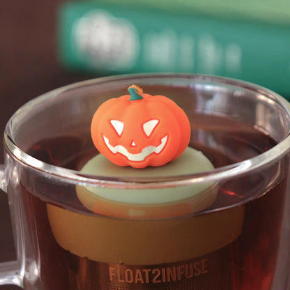 Jack-o-lantern pumpkin tea infuser for loose leaf tea  with a fine mesh stainless steel tea strainer that reads FLOAT2INFUSE on a green base floating in a glass tea mug with books in the background