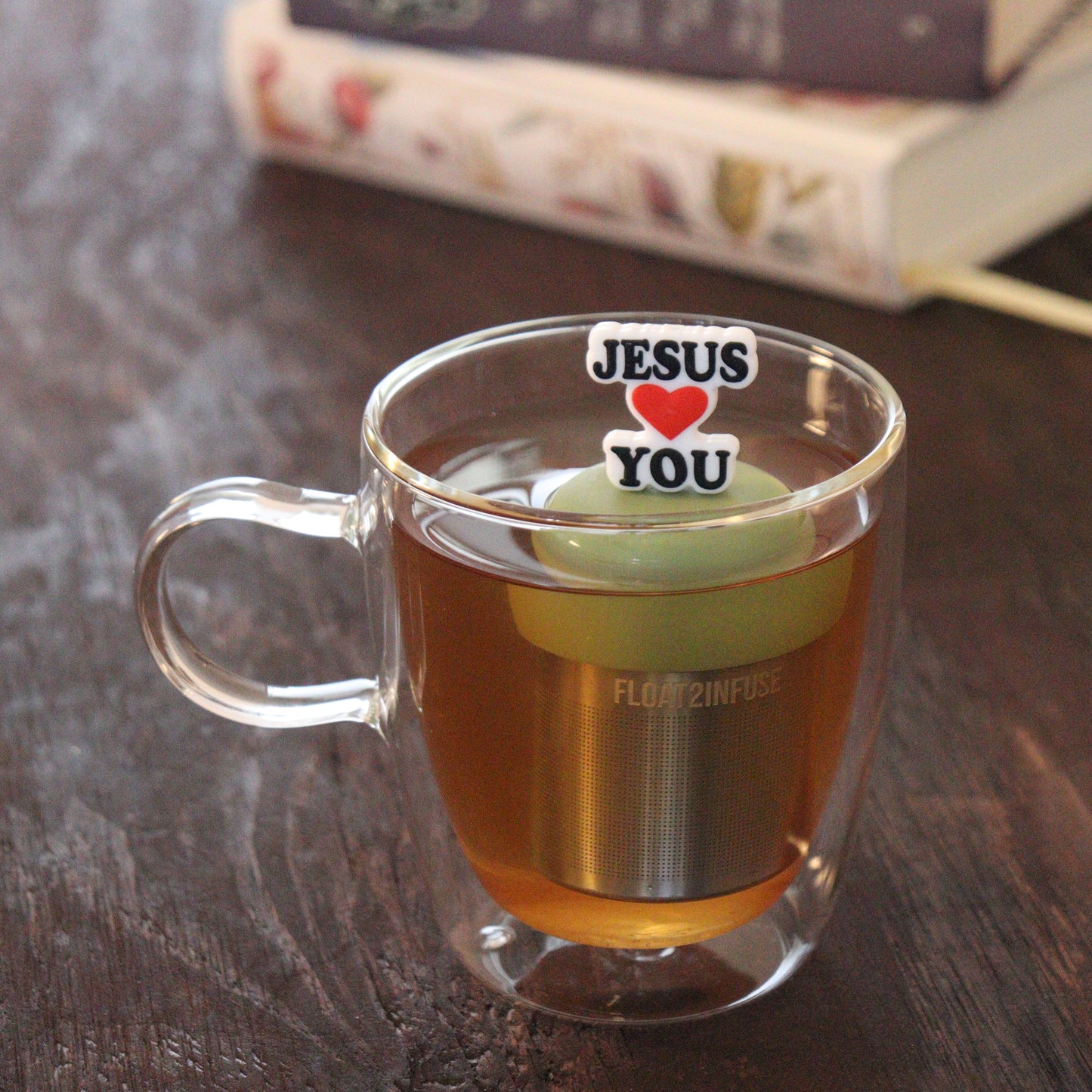 black text with red heart that reads "jesus loves you" tea infuser for loose leaf tea  with a fine mesh stainless steel tea infuser that reads FLOAT2INFUSE on a green base floating in a glass tea mug with books in the background