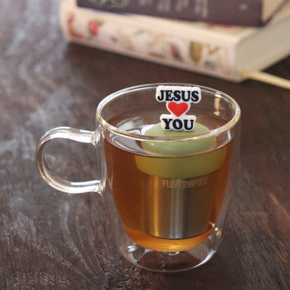 black text with red heart that reads "jesus loves you" tea infuser for loose leaf tea  with a fine mesh stainless steel tea infuser that reads FLOAT2INFUSE on a green base floating in a glass tea mug with books in the background