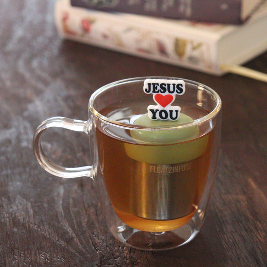 black text with red heart that reads "jesus loves you" tea infuser for loose leaf tea  with a fine mesh stainless steel tea infuser that reads FLOAT2INFUSE on a green base floating in a glass tea mug with books in the background