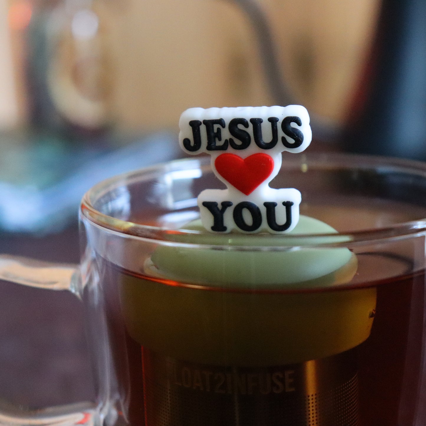"jesus loves you" text tea infuser for loose leaf tea  with a fine mesh stainless steel tea strainer that reads FLOAT2INFUSE on a green base floating in a glass tea mug with tea kettle in the background