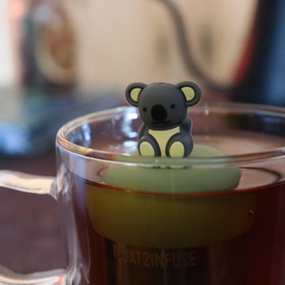 grey koala animal tea infuser for loose leaf tea with a fine mesh stainless steel tea strainer that reads FLOAT2INFUSE on a green base  floating in a glass tea mug with tea kettle in the background