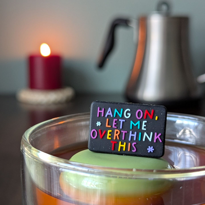 black rectangle with text that reads "hang on, let me overthink this" in rainbow letters tea infuser  for loose leaf tea with fine mesh stainless steel tea strainer that reads FLOAT2INFUSE on a green base with a lit candle and a tea kettle in the background