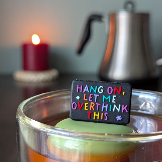black rectangle with text that reads "hang on, let me overthink this" in rainbow letters tea infuser  for loose leaf tea with fine mesh stainless steel tea strainer that reads FLOAT2INFUSE on a green base with a lit candle and a tea kettle in the background
