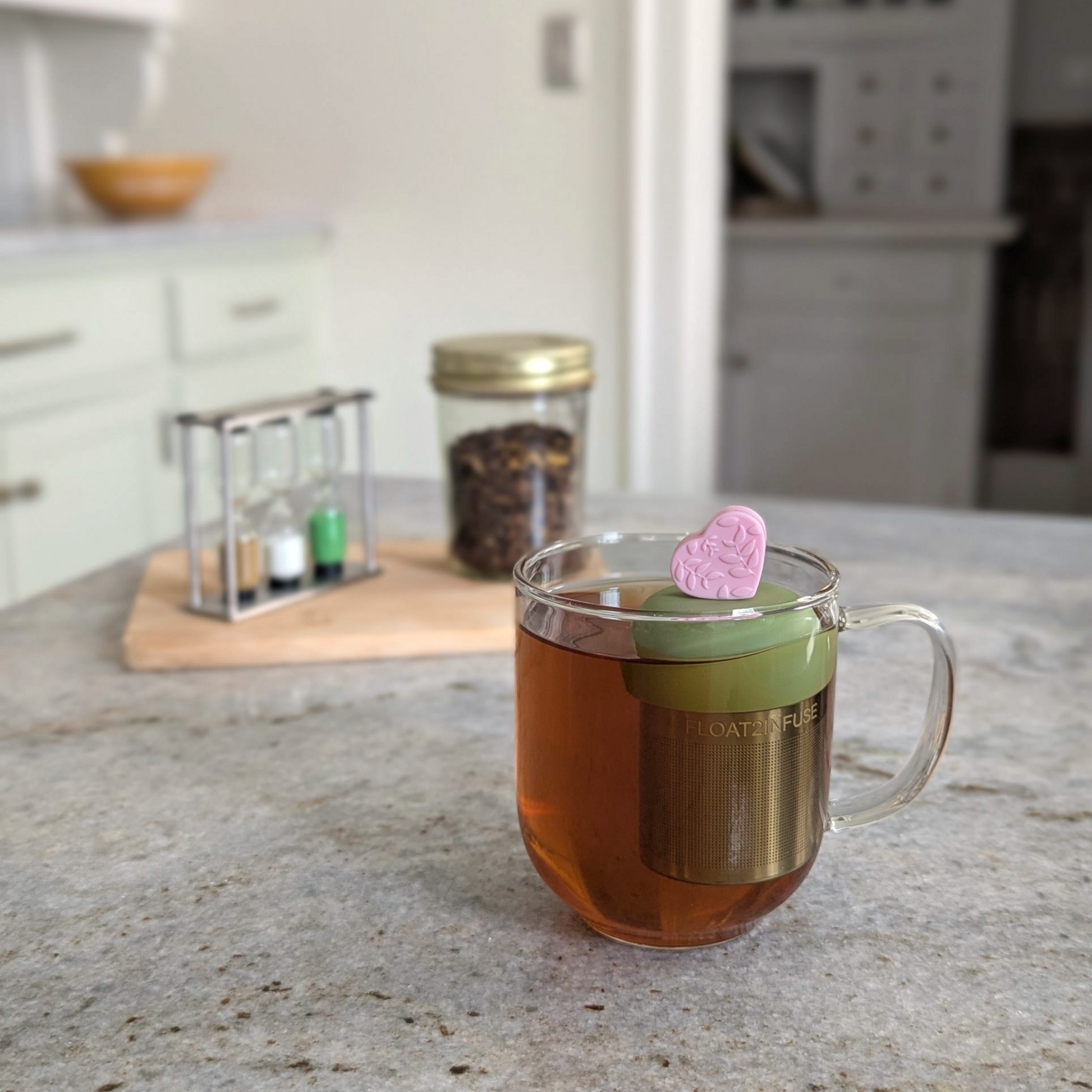 pink heart with a floral leaf pattern tea infuser  for loose leaf tea with fine mesh stainless steel tea strainer that reads FLOAT2INFUSE on a green base floating in a glass tea mug with loose leaf tea in the background