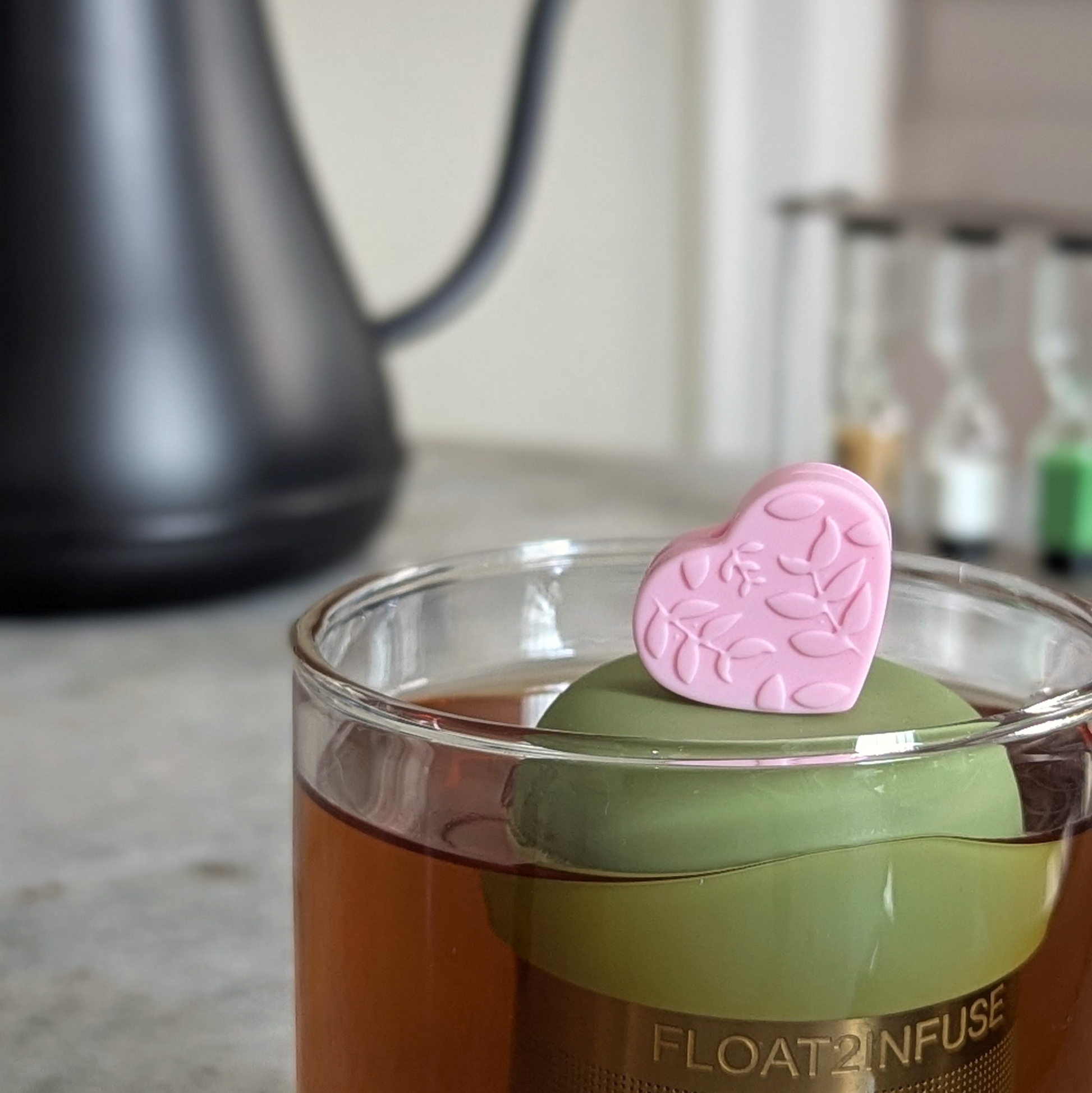 pink heart with a floral leaf pattern tea infuser  for loose leaf tea with fine mesh stainless steel tea strainer that reads FLOAT2INFUSE on a green base floating in a glass tea mug with loose leaf tea in the background
