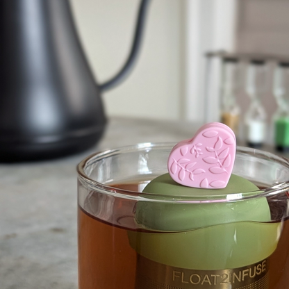 pink heart with a floral leaf pattern tea infuser  for loose leaf tea with fine mesh stainless steel tea strainer that reads FLOAT2INFUSE on a green base floating in a glass tea mug with loose leaf tea in the background