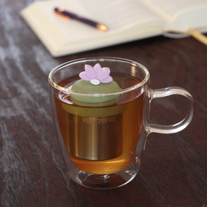 light pink lotus flower tea infuser for loose leaf tea  with a fine mesh stainless steel tea infuser that reads FLOAT2INFUSE on a green base floating in a glass tea mug with notebook and pen in the background