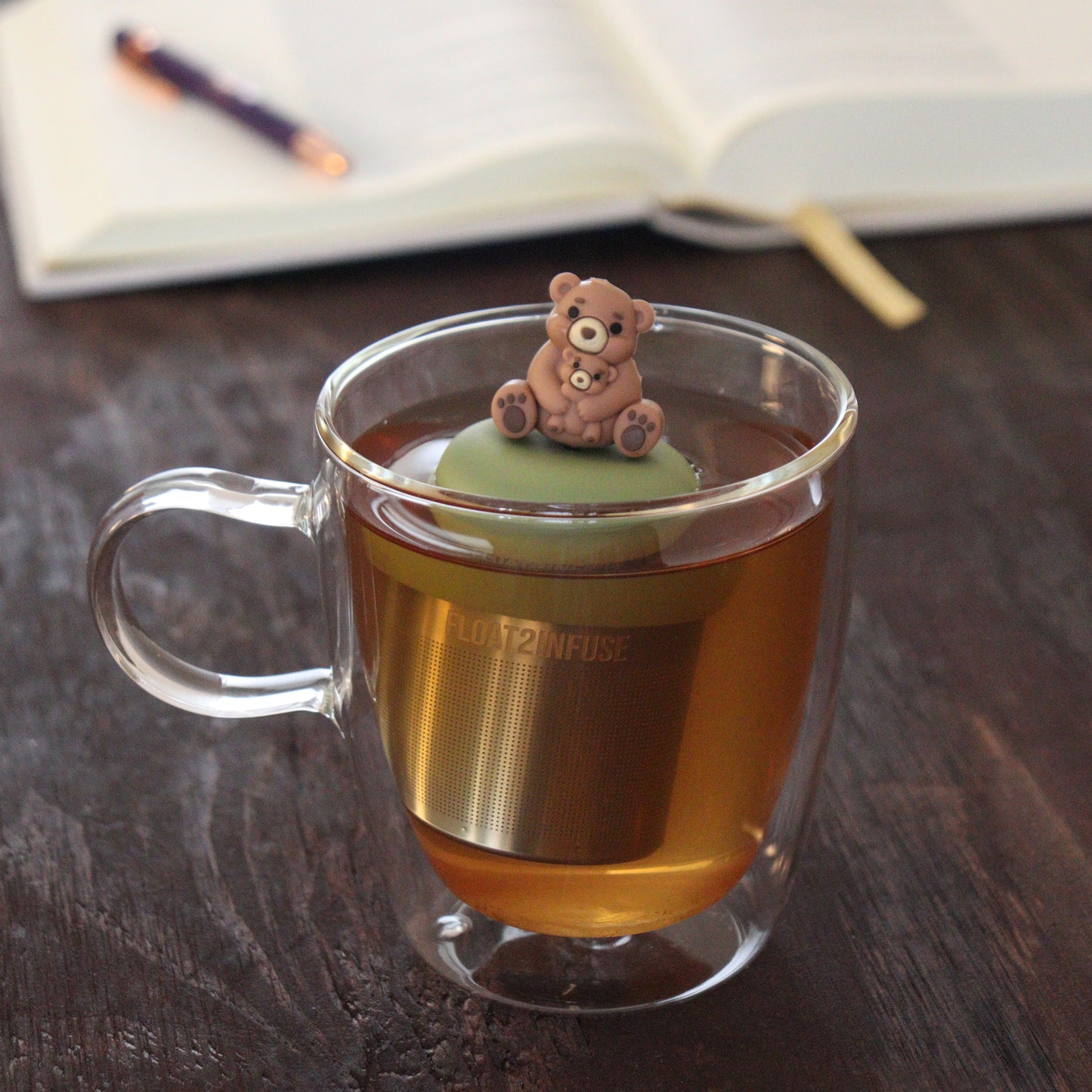 brown bear mom and baby bear animal tea infuser for loose leaf tea with a fine mesh stainless steel tea infuser that reads FLOAT2INFUSE on a green base floating in a glass tea mug with notebook and pen in the background