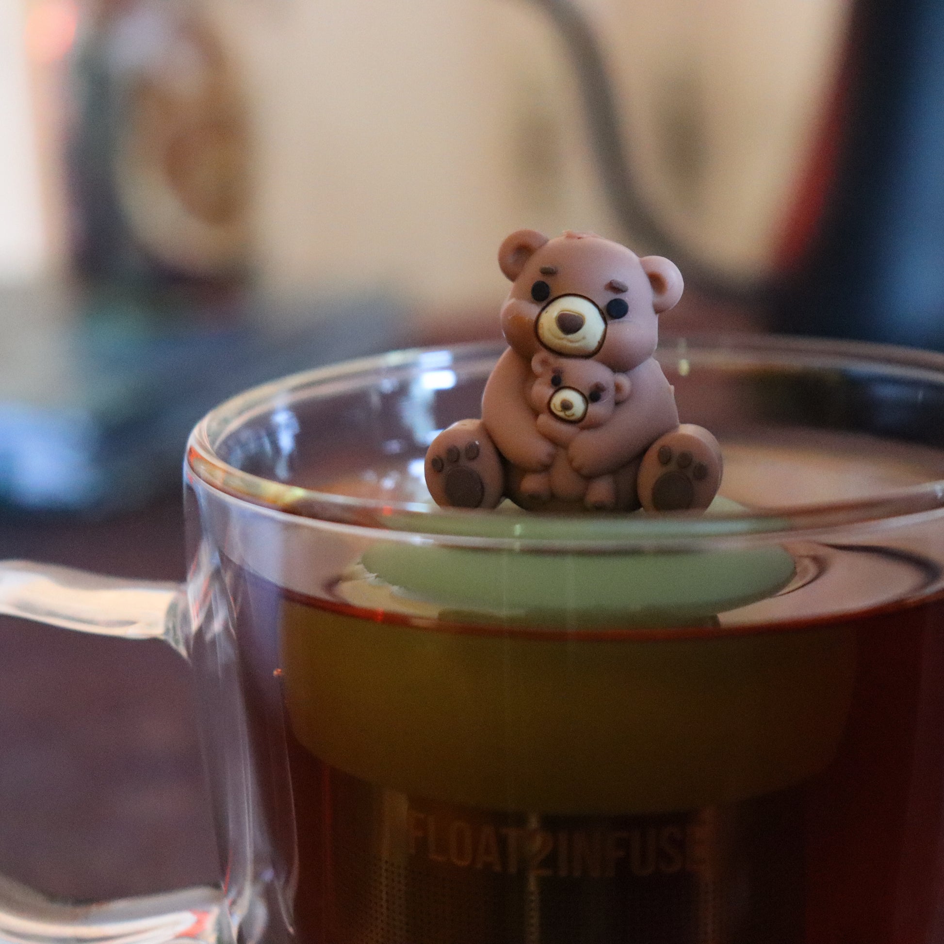 brown mom bear and baby bear animal tea infuser for loose leaf tea  with a fine mesh stainless steel tea strainer that reads FLOAT2INFUSE on a green base floating in a glass tea mug with tea kettle in the background